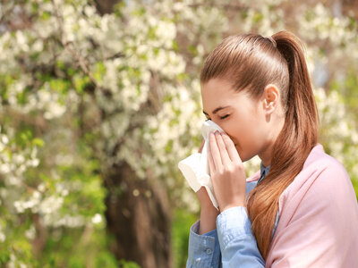 Picture of a female standing outside near blossomed flowers in the spring time. She sneezing into a kleenex.